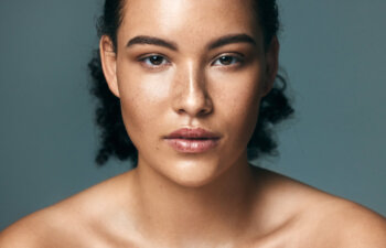A young woman with curly hair and freckles looks directly at the camera against a plain, neutral background. She has a calm, neutral expression and bare shoulders.