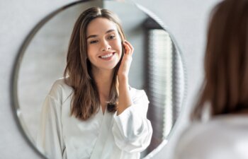 Beauty Concept. Portrait Of Attractive Happy Woman Looking At Mirror In Bathroom, Beautiful Millennial Lady Wearing White Silk Robe Smiling To Reflection, Enjoying Her Appearance, Selective Focus